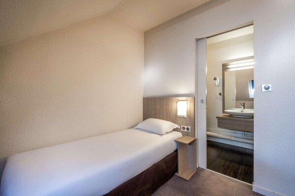 A small, neatly made bed in a hotel room with a wooden headboard, a bedside table with a lamp, and a mirror above the sink.