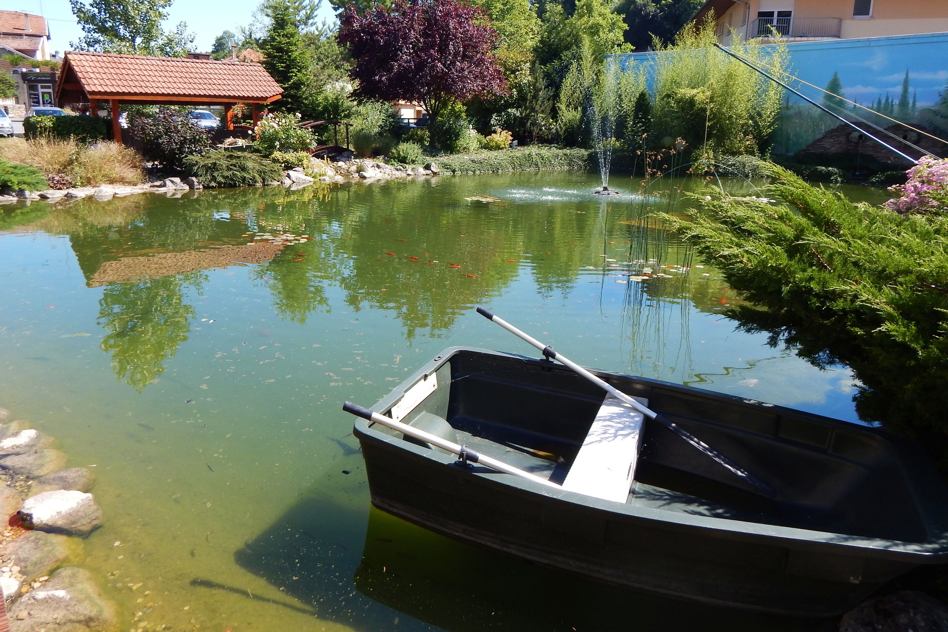 Ein kleines Boot liegt an einem ruhigen Teich mit einem Brunnen, umgeben von üppiger Vegetation und einem Gebäude mit rotem Dach.