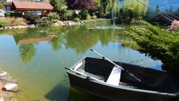 Ein kleines Boot liegt an einem ruhigen Teich mit einem Brunnen, umgeben von üppiger Vegetation und einem Gebäude mit rotem Dach.