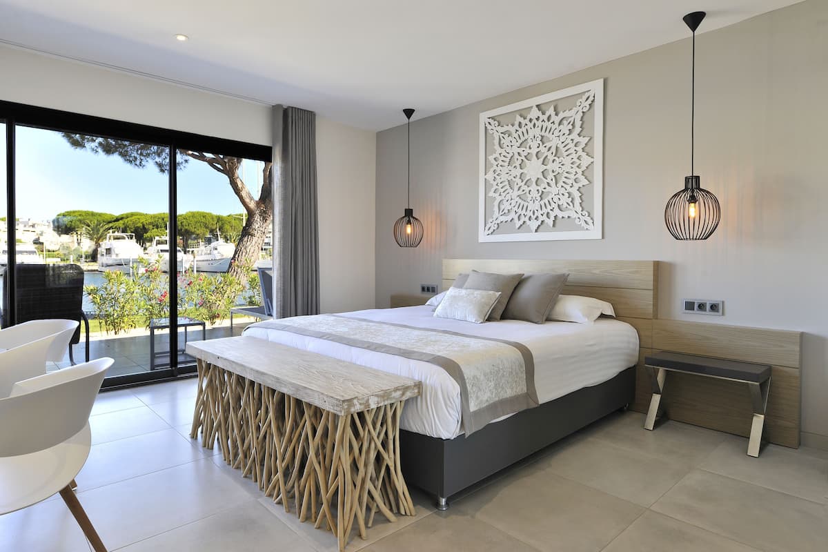 A modern bedroom with a large bed, a bench, a table, and a view of the marina.