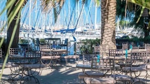 An outdoor seating area with tables and chairs, a palm tree, and a marina with boats in the background. - Le Spinaker Boutique Hôtel Restaurant (Le Grau-Du-Roi)