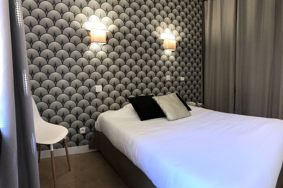 Habitación de hotel con papel tapiz estampado, cama con ropa de cama blanca, dos luces fijadas a la pared y una puerta que da al pasillo.