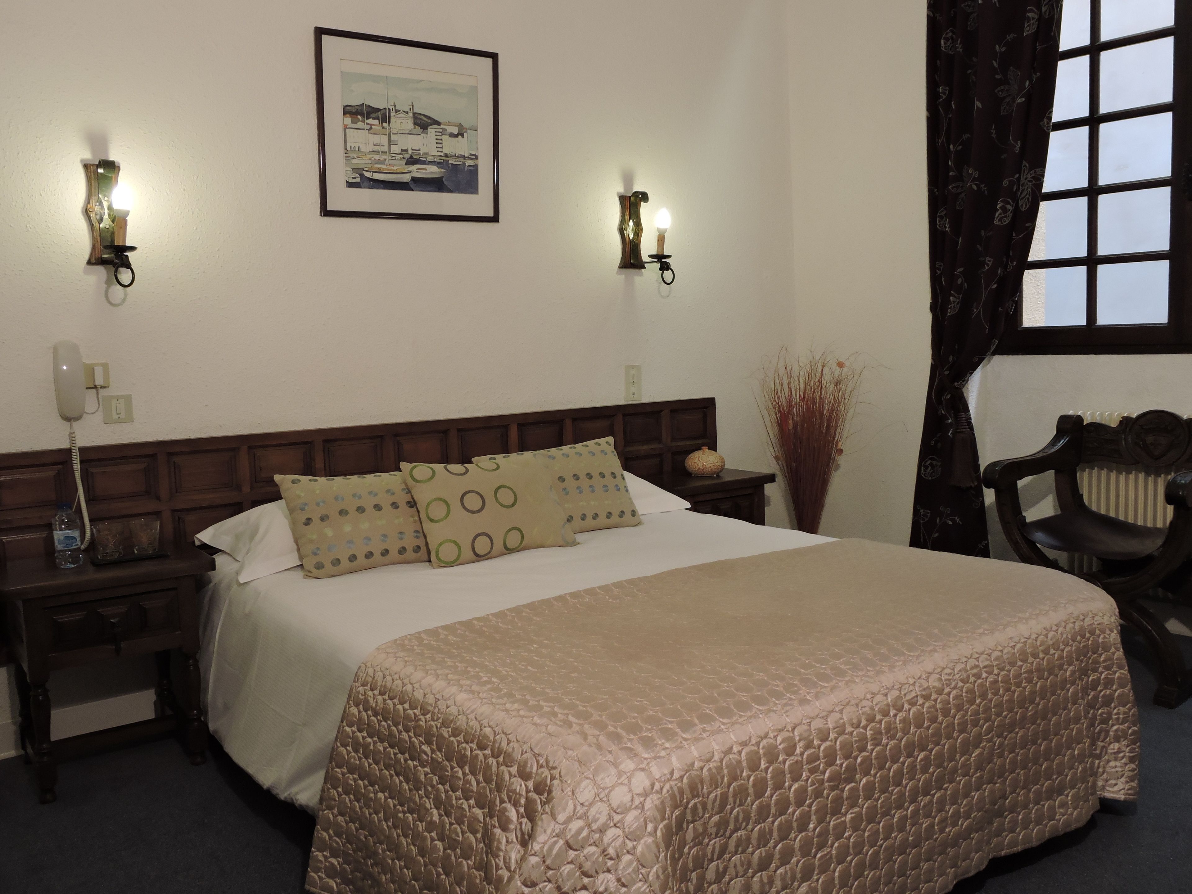 A neatly made bed with a patterned bedspread, two bedside tables with lamps, a framed artwork on the wall, and a window with curtains.