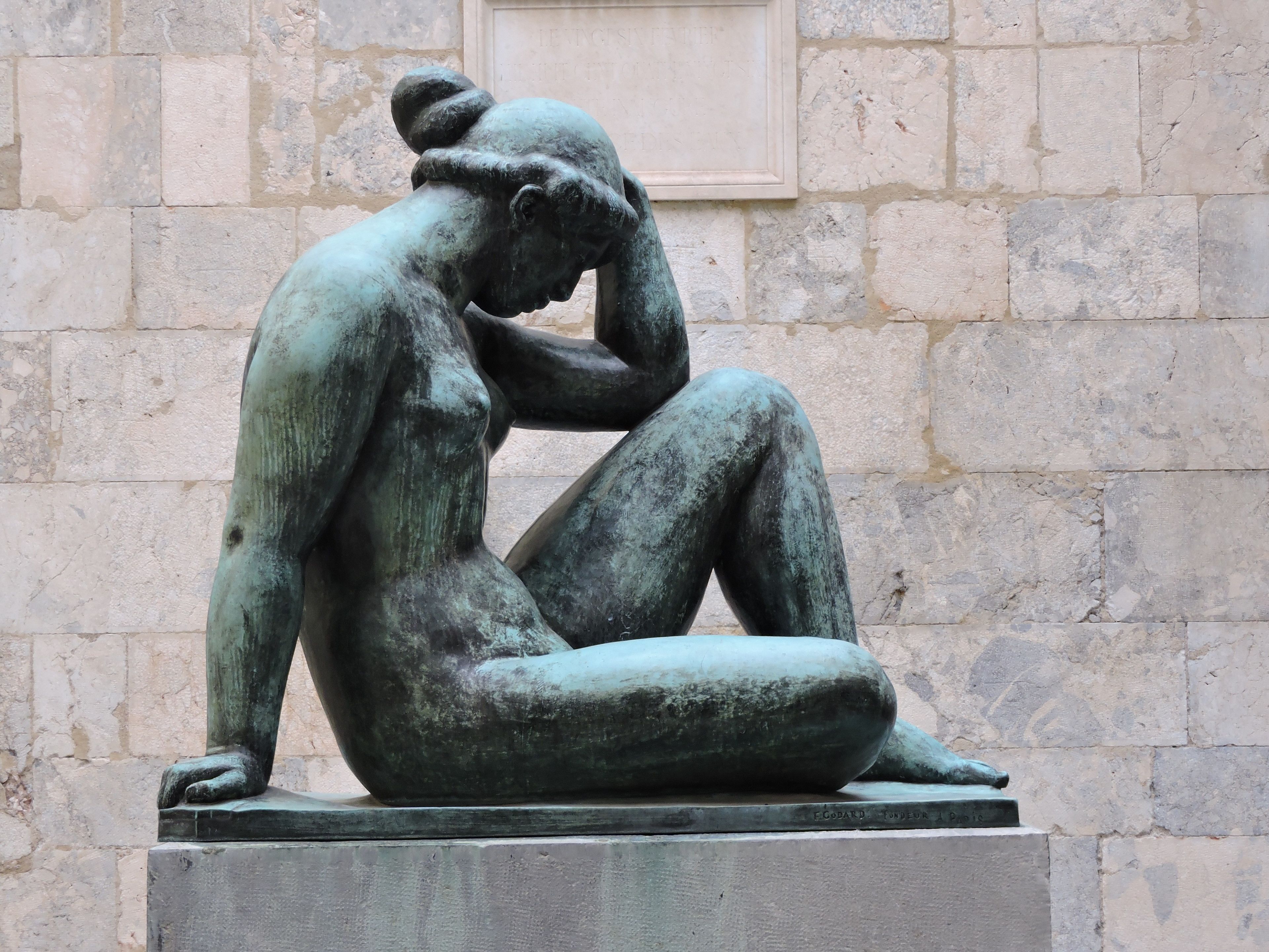Una escultura de bronce de una figura sentada en posición contemplativa, sobre un fondo de pared de piedra.