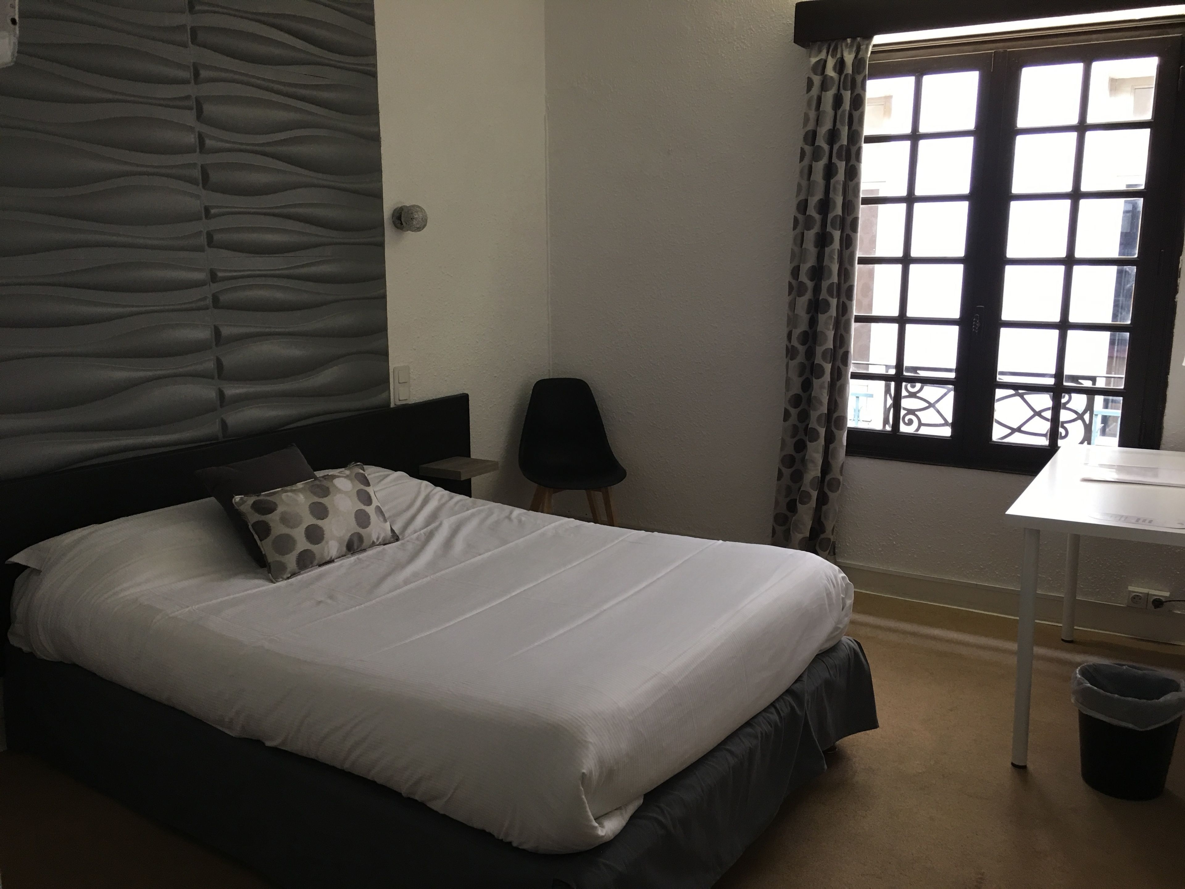 Una cama bien hecha con cabecera, una mesita de noche pequeña, una silla y una ventana con cortinas.