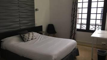 Una cama bien hecha con cabecera, una mesita de noche pequeña, una silla y una ventana con cortinas.
