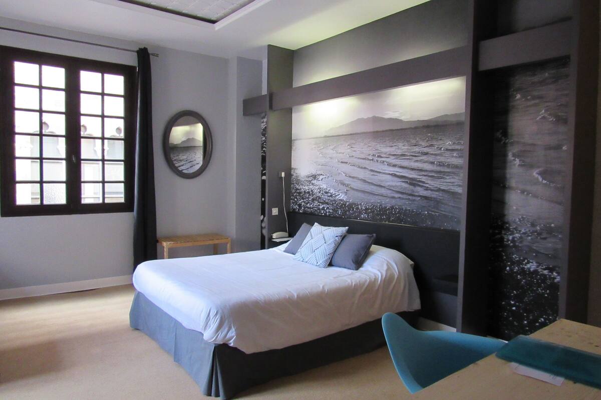 Un dormitorio moderno con una cama grande, una mesita de noche, un espejo y una obra de arte en la pared que representa una playa.