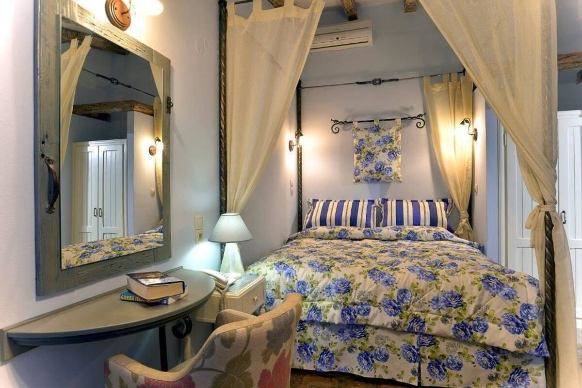 A bedroom with a bed, a bedside table with a lamp, a chair, a mirror, and a window with curtains.