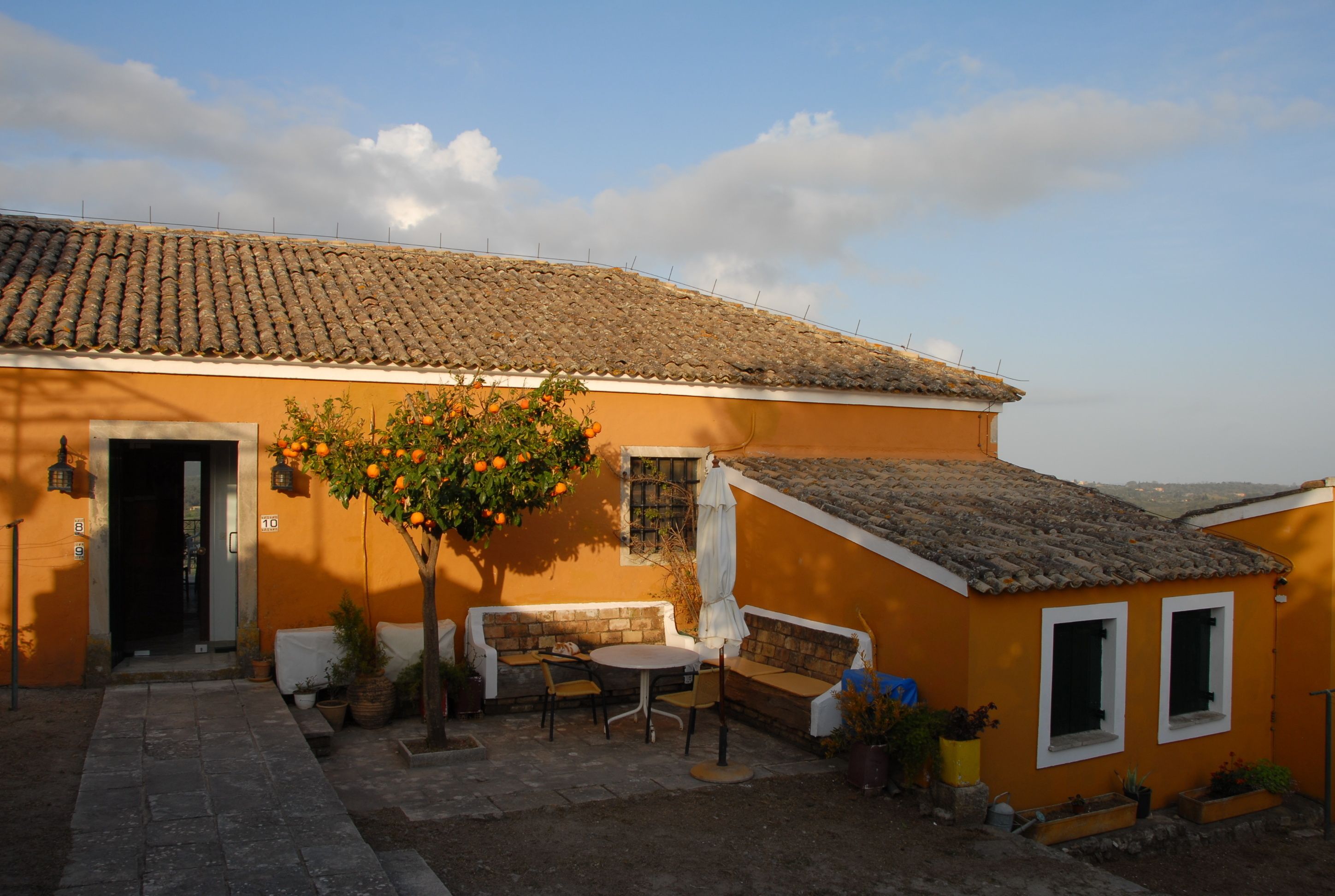 A building with a tiled roof, a small patio with a table and chairs, and a tree with orange fruit.