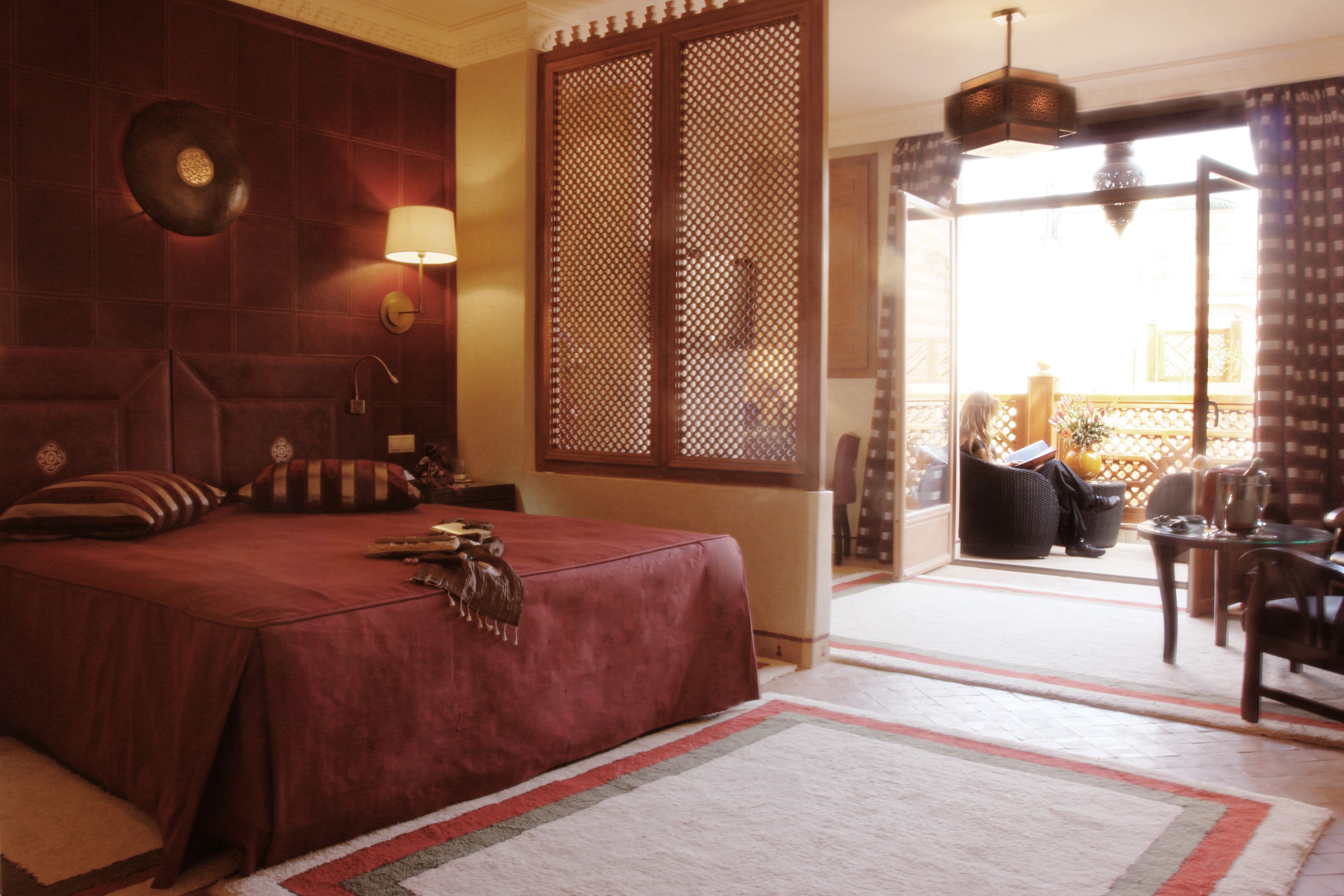 Una habitación de hotel con una cama grande, una zona de estar con una mesa y sillas, vista al balcón exterior y una pared decorativa con una lámpara.