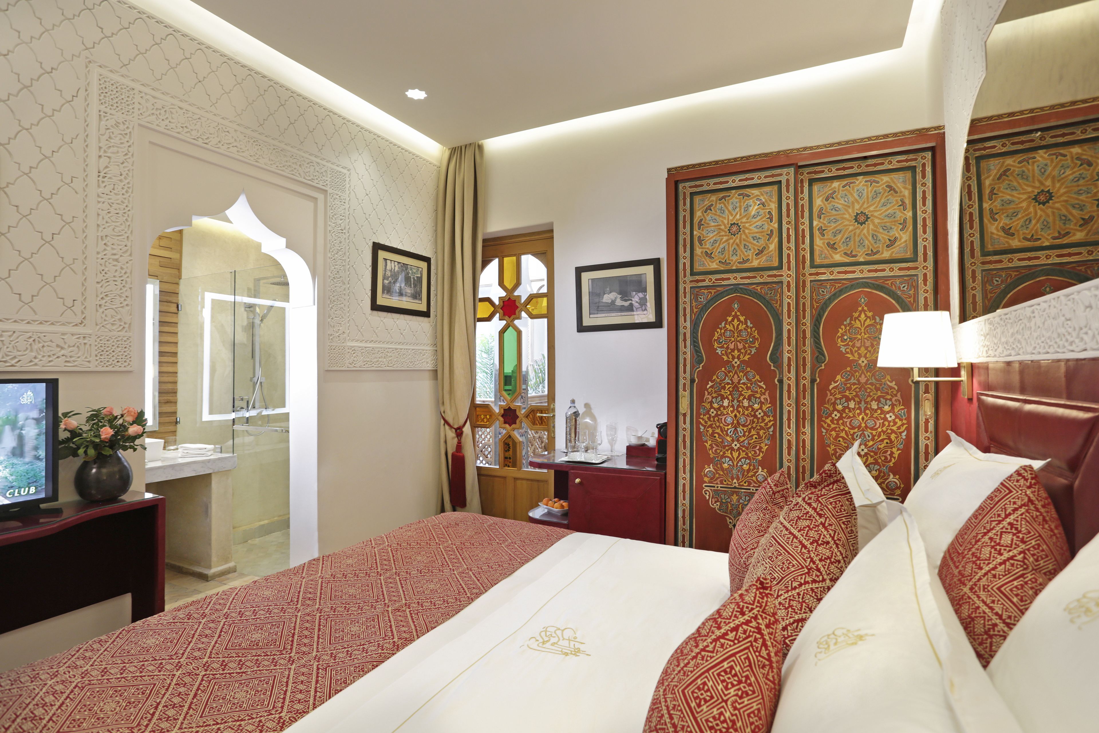 Habitación de hotel con una cama grande, televisión, escritorio con un florero y baño visible a través de un arco.