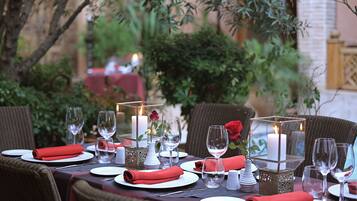 Un espacio de comedor al aire libre con mesas dispuestas para dos personas, con platos blancos, servilletas rojas y copas de vidrio transparente.