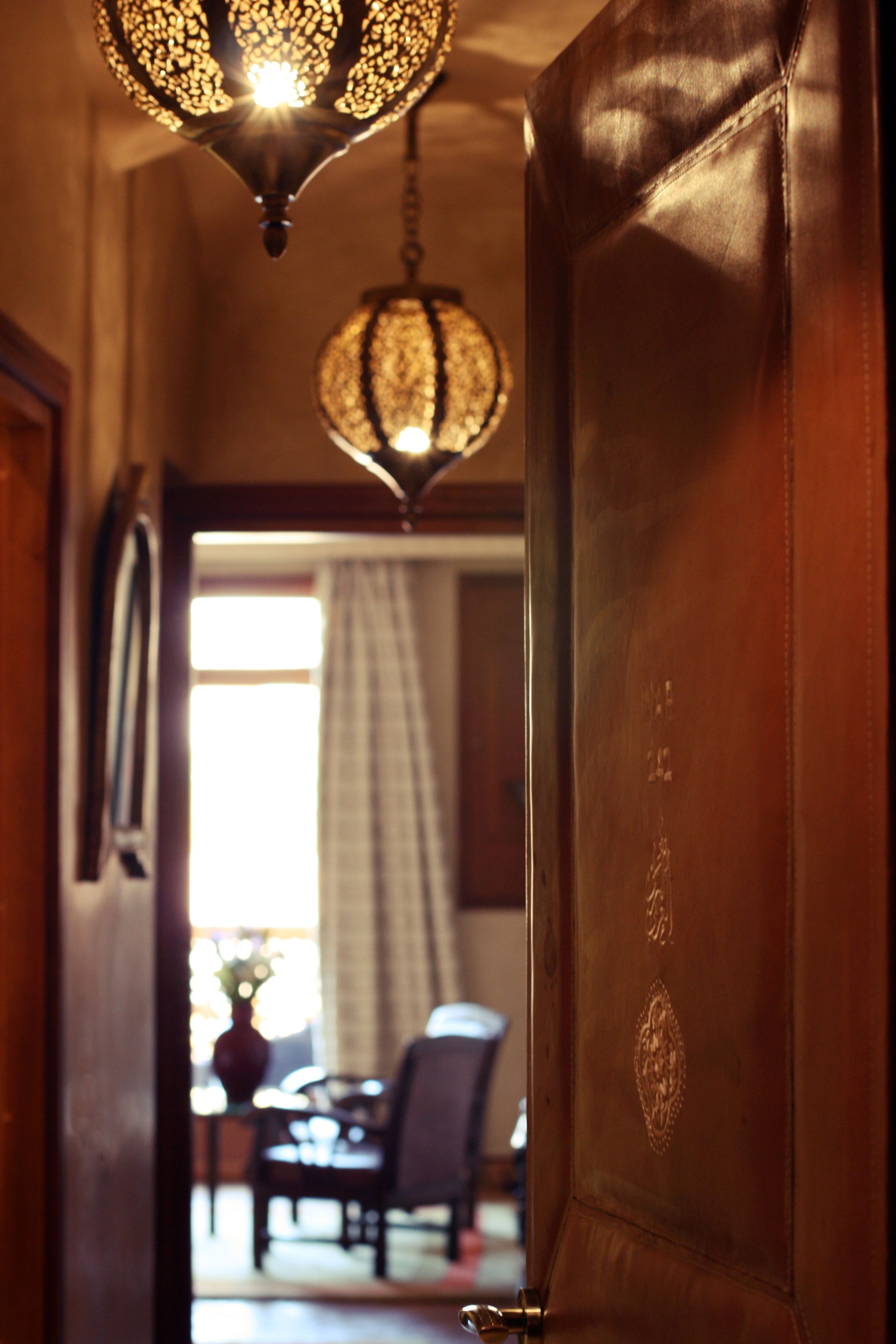 Una puerta de madera con manija de latón, que da a una habitación con una mesa de comedor y sillas, y una lámpara colgante con pantalla decorada.