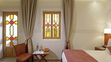 Habitación de hotel con una cama, una silla, una mesita con una bandeja de naranjas, una botella y vasos.