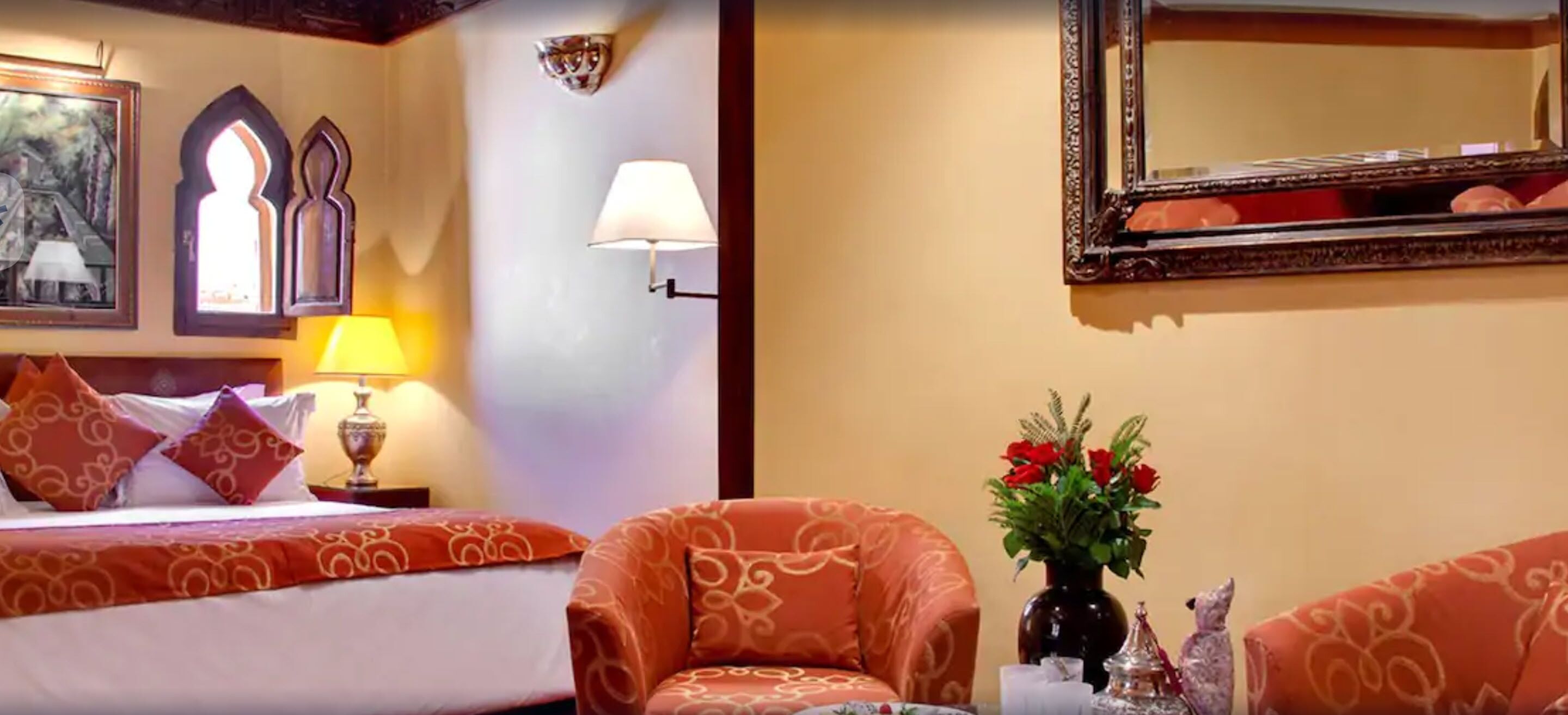 A hotel room with a bed, two chairs, a lamp, a vase with flowers, and a mirror on the wall.