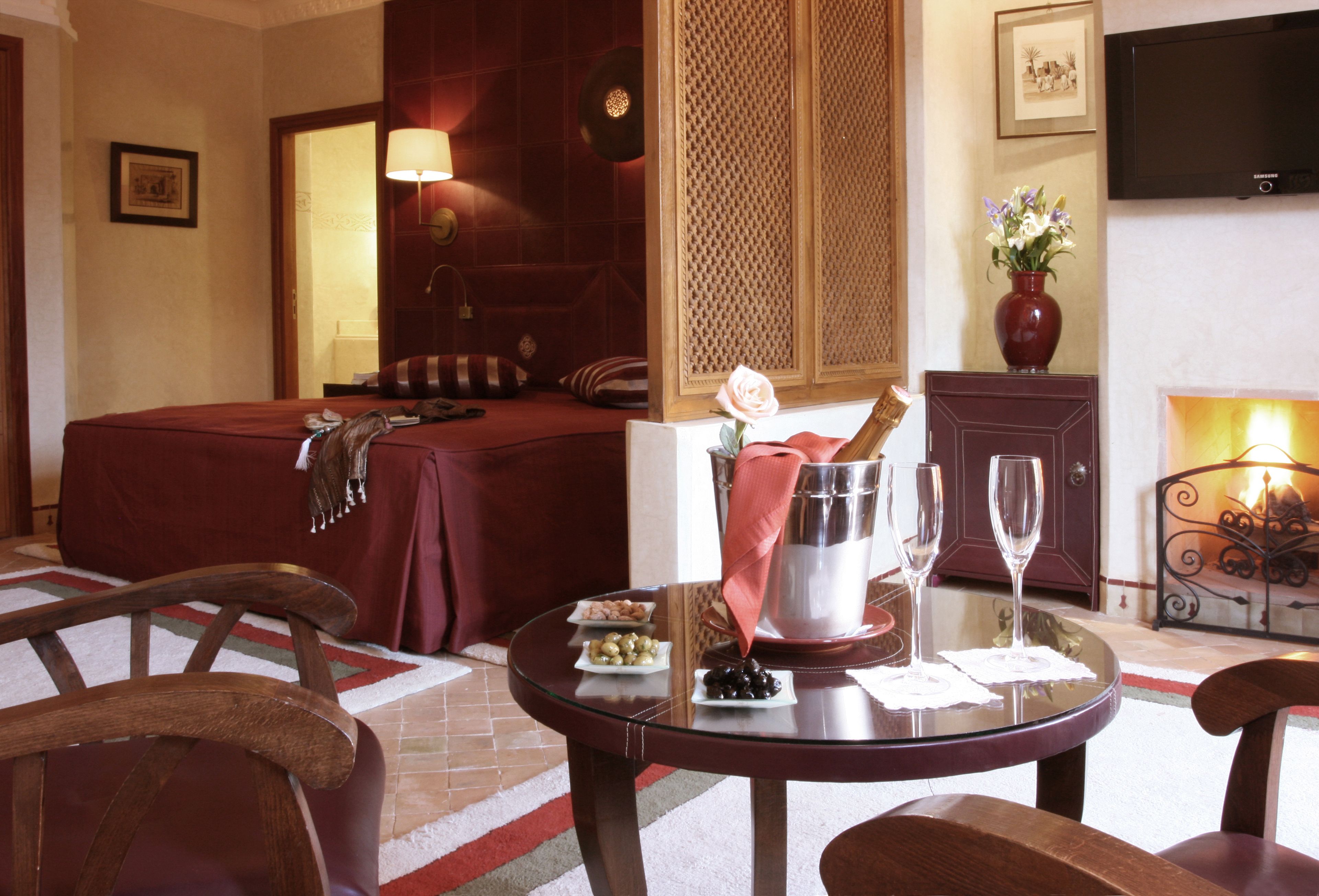 A hotel room with a bed, a dining table with a vase of flowers, a wine glass, and a minibar.