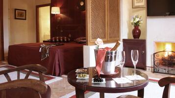 A hotel room with a bed, a dining table with a vase of flowers, a wine glass, and a minibar.