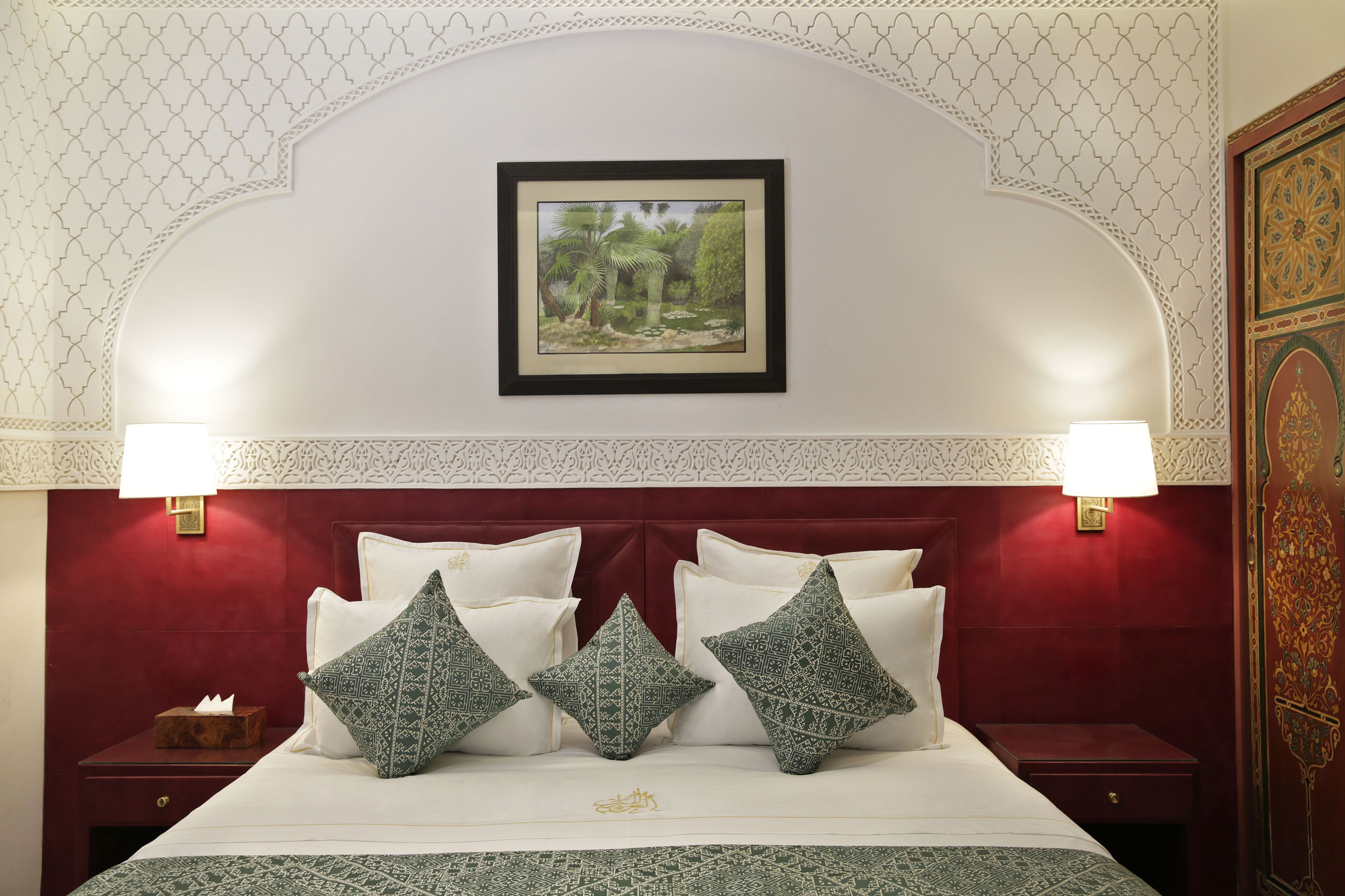A hotel room with a red headboard, white bedding, and decorative pillows. There is a framed picture on the wall above the bed and two wall-mounted lamps on either side.