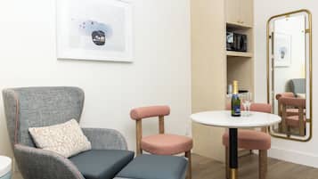 A modern room with a grey armchair, a small round table with a bottle and glass, and a mirror with a gold frame.