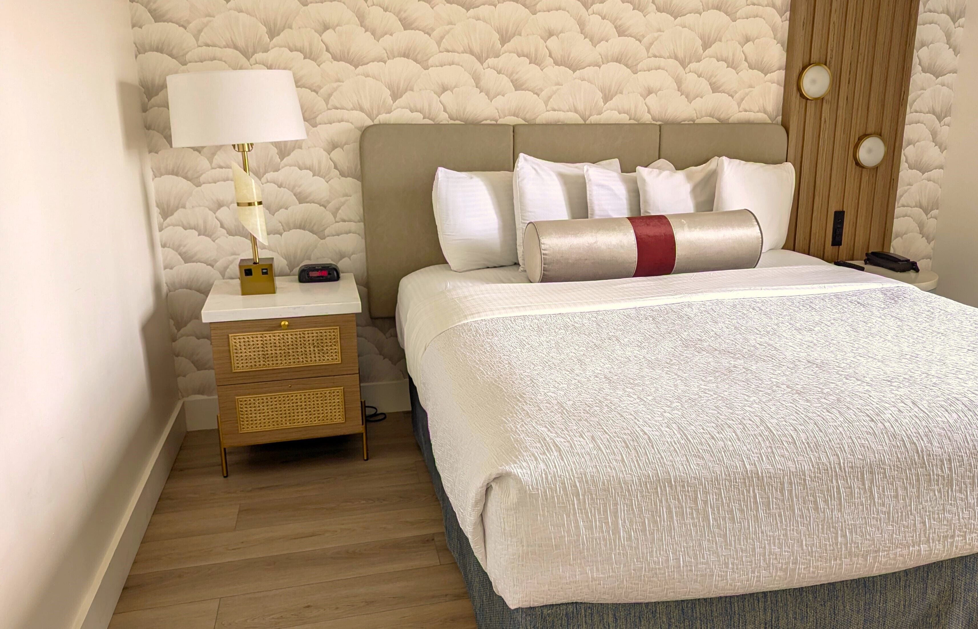 Quarto de hotel com cama, mesa de cabeceira com luminária, mesa de apoio de madeira e parede com padrão texturizado.