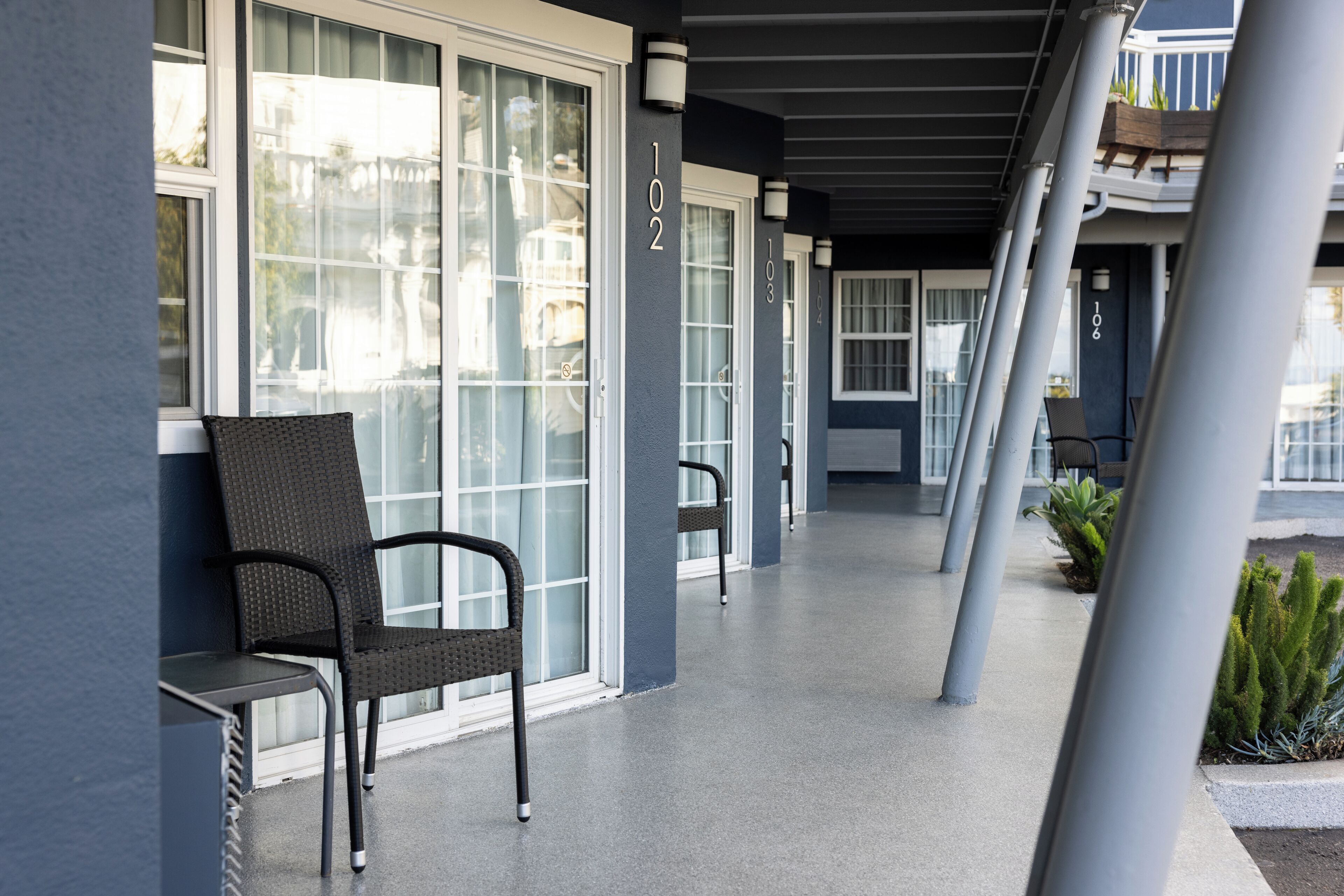 A covered outdoor area with chairs and potted plants, featuring large windows and a number '102' on the wall.