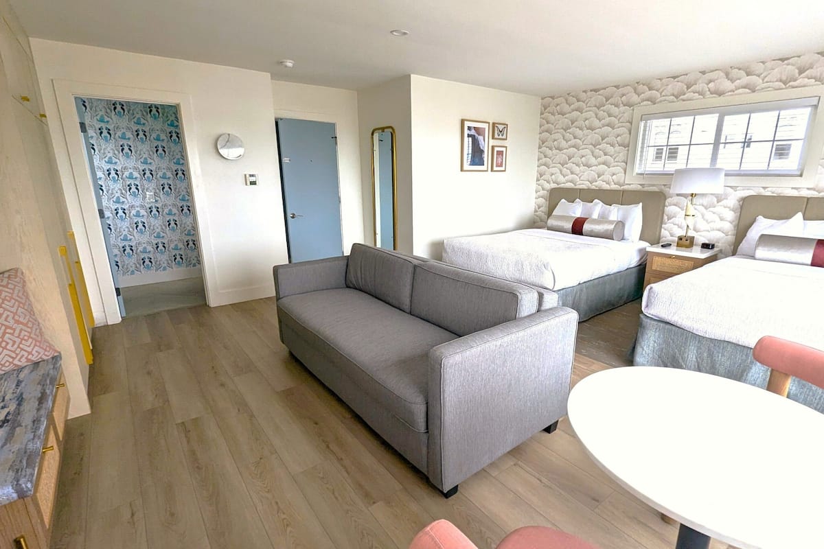 Quarto de hotel com sofá, duas camas, uma mesinha e um detalhe decorativo na parede.