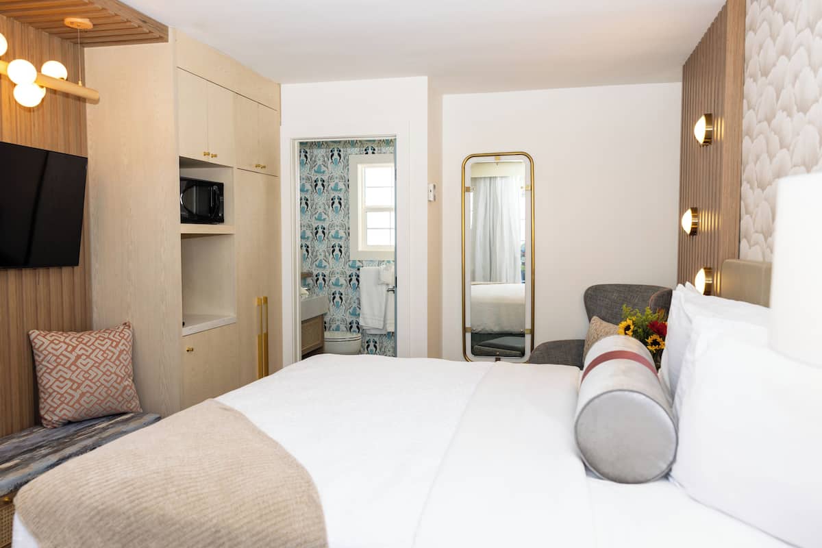 Quarto de hotel com uma cama, televisão, guarda-roupa, espelho e banheiro visível por uma porta aberta.