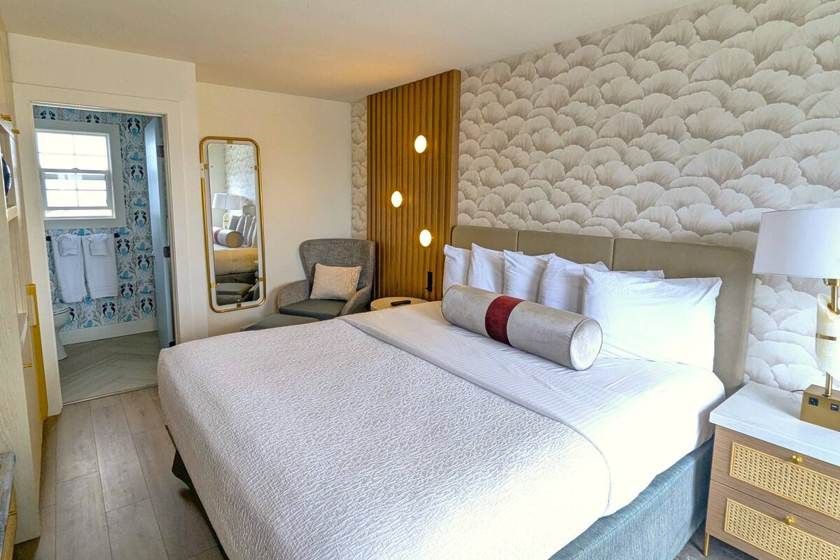 Quarto de hotel com uma cama grande, uma mesa de cabeceira com luminária, uma cadeira, um espelho e banheiro com chuveiro e vaso sanitário visíveis através de uma porta aberta.