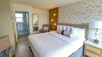 Habitación de hotel con una cama grande, una mesita de noche con lámpara, una silla, un espejo y un baño con ducha e inodoro visibles a través de una puerta abierta.