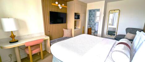 A hotel room with a bed, a desk with a lamp, a television, a bathroom, and a mirror.