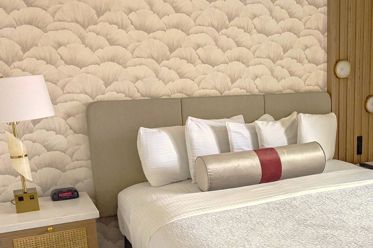 Quarto de hotel com cama, mesa de cabeceira, luminária e parede com textura.
