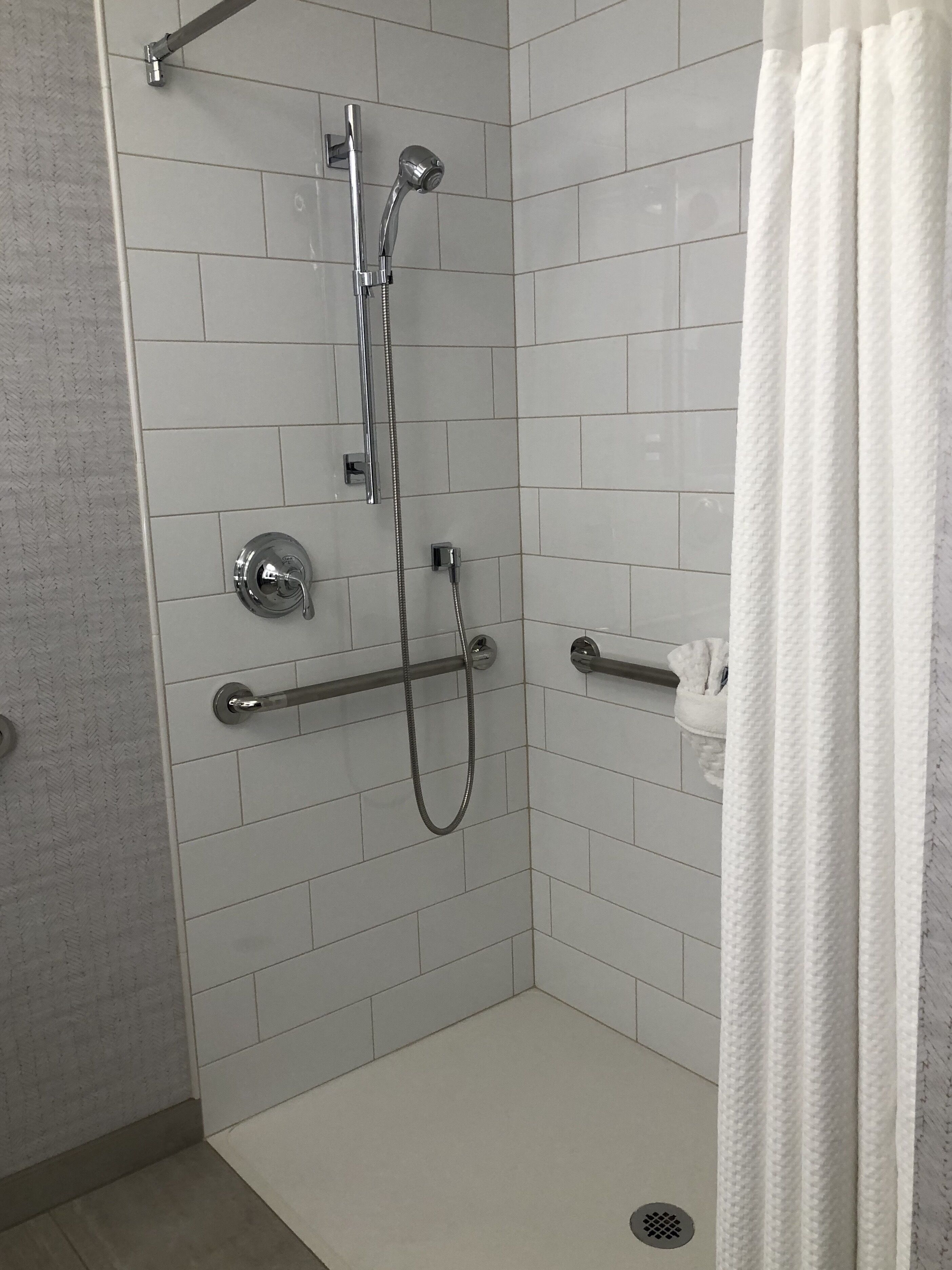 Ducha a ras de suelo con paredes de azulejos blancos, una barra de apoyo y un cabezal de ducha.