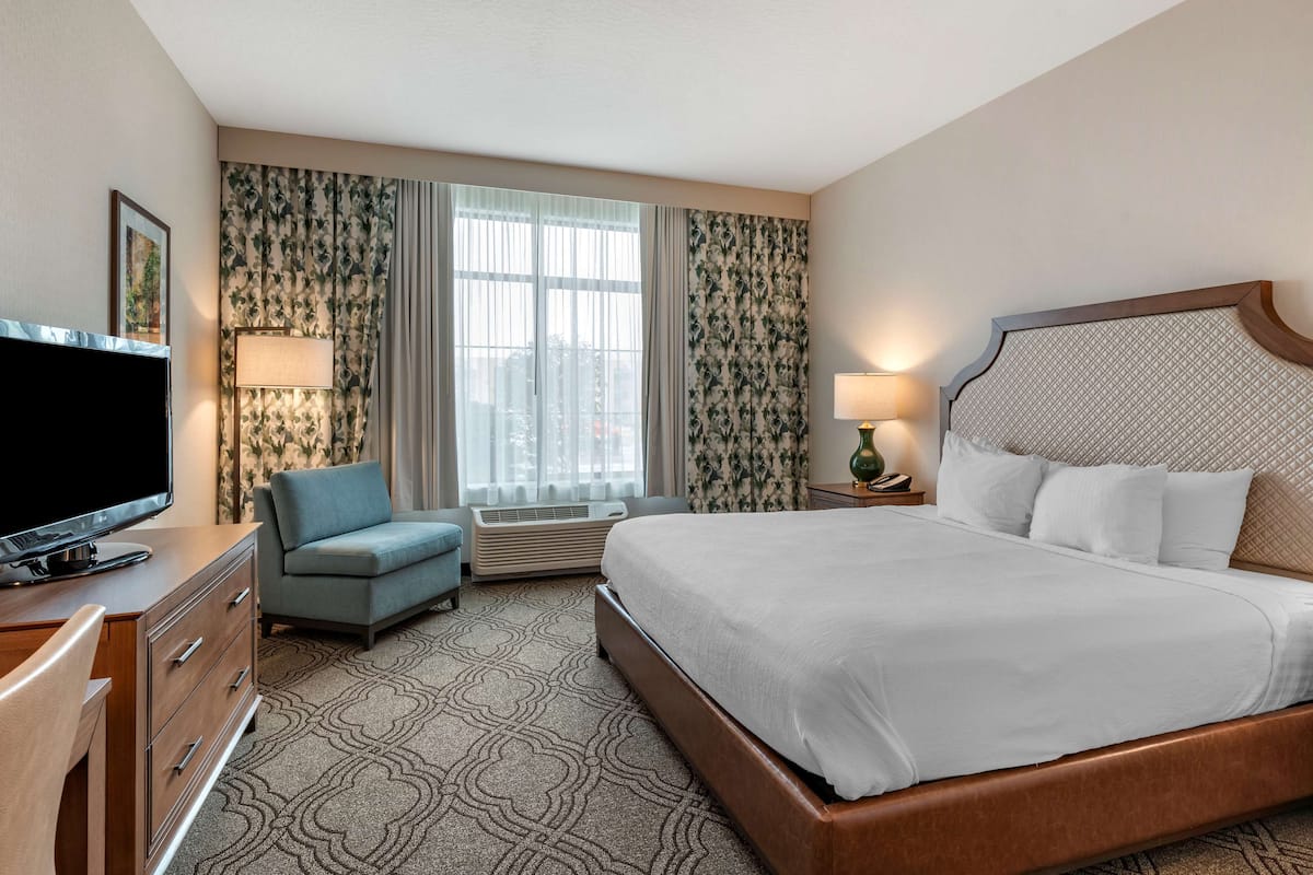 A hotel room with a large bed, a television, a desk with drawers, a chair, a lamp, and curtains.
