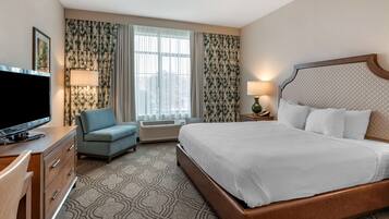 Habitación de hotel con una cama grande, televisión, escritorio con cajones, una silla, lámpara y cortinas.
