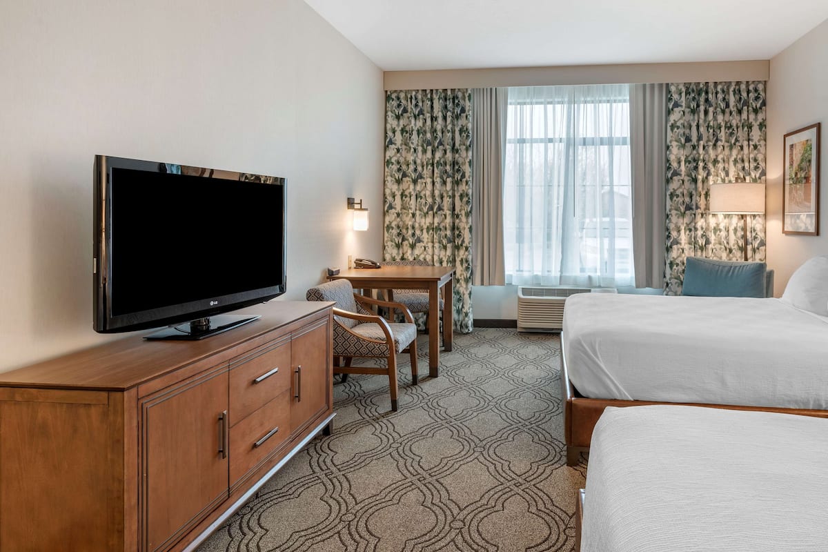 A hotel room with a TV on a wooden cabinet, a desk with a chair, two beds, and a window with curtains.