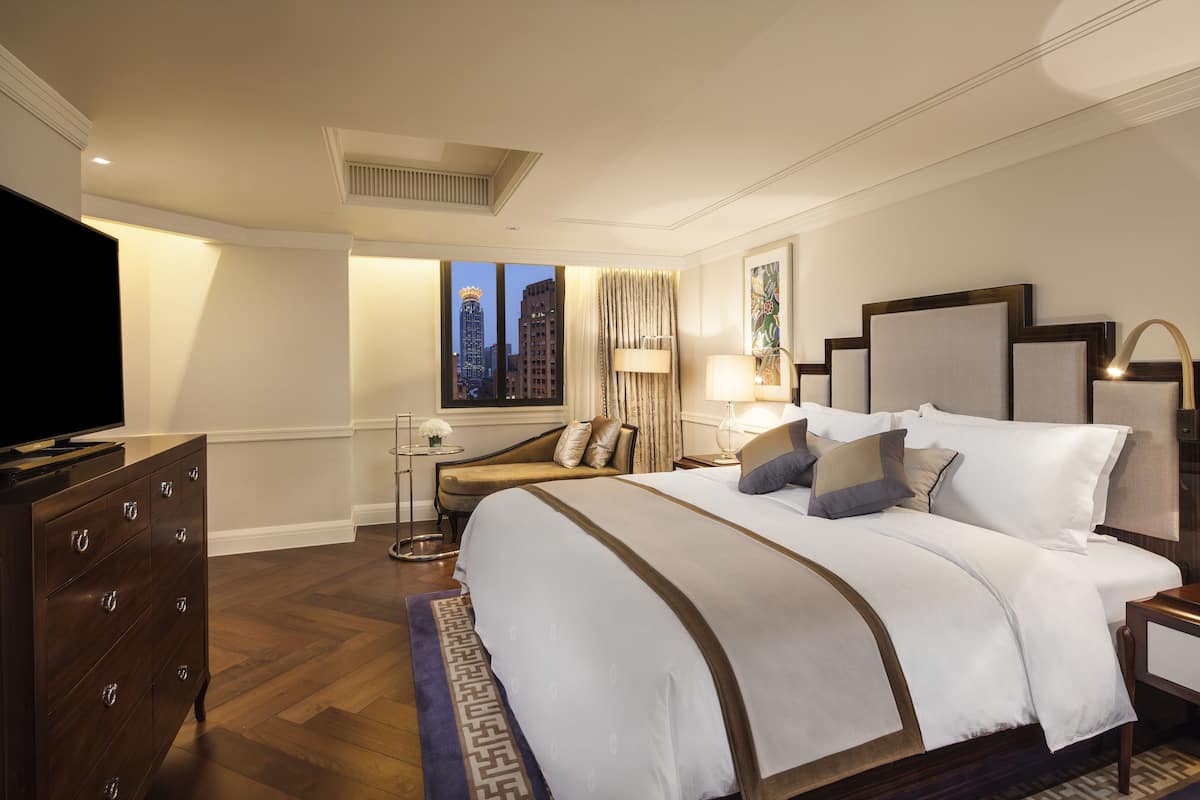A hotel room with a large bed, a dresser, a television, a nightstand, and a window with a city view.