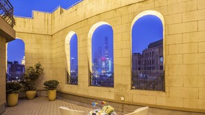 Une terrasse extérieure avec une table et des chaises, des plantes en pot et de grandes fenêtres en arc d’où l’on peut admirer la vue sur la ville au crépuscule.