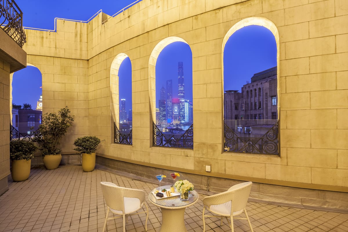 An outdoor terrace with a table and chairs, potted plants, and large arched windows overlooking a cityscape at dusk.