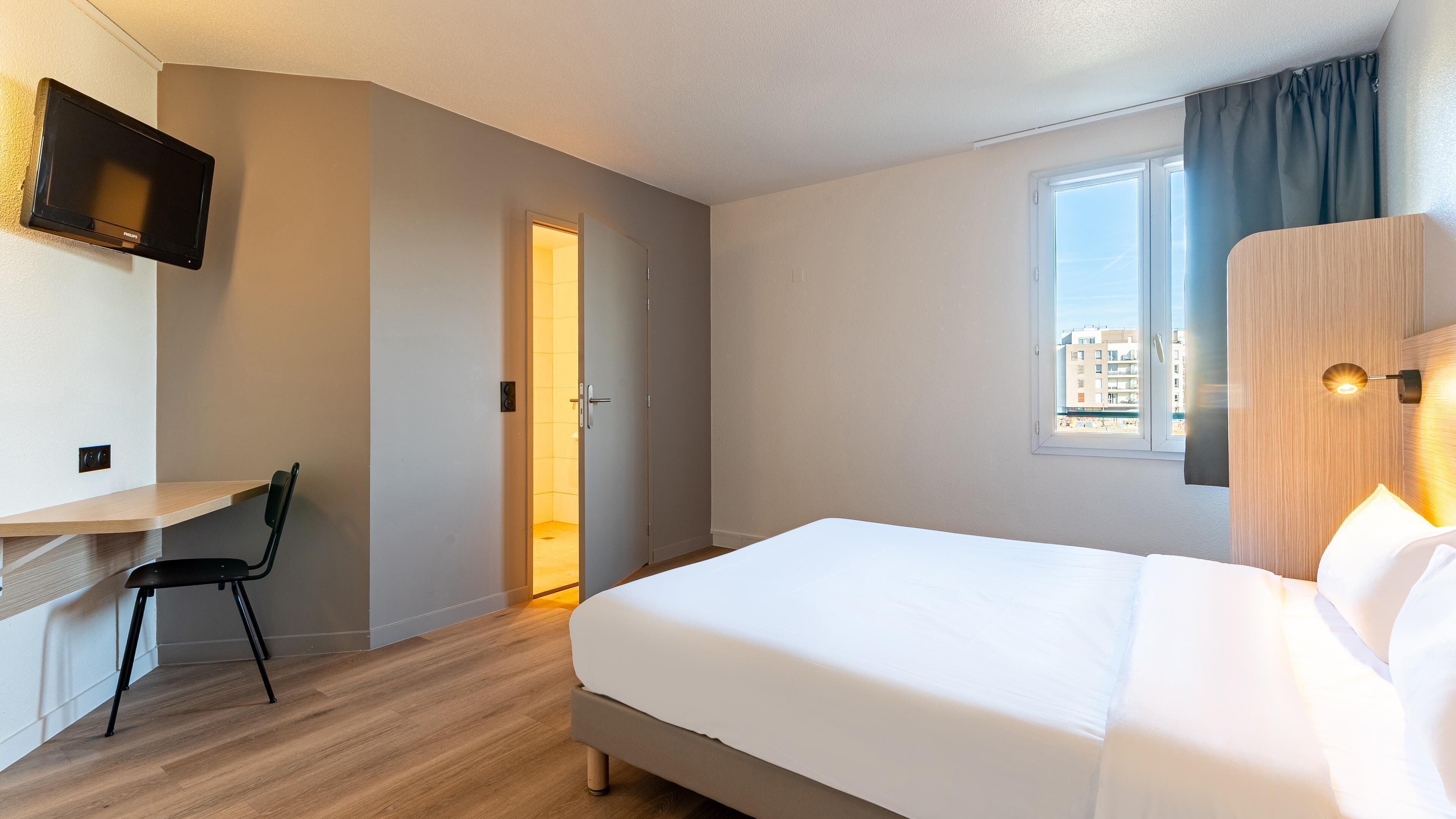 A hotel room with a bed, a desk with a chair, a TV mounted on the wall, a window with curtains, and a view of buildings outside.