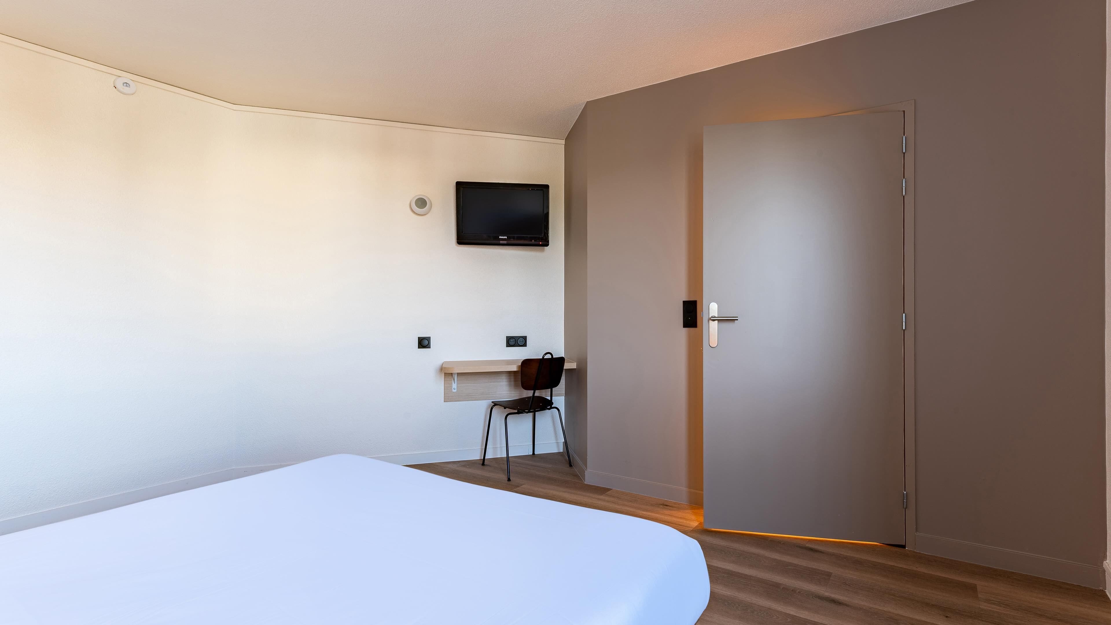 A hotel room with a bed, a desk, a chair, a television mounted on the wall, and a door.