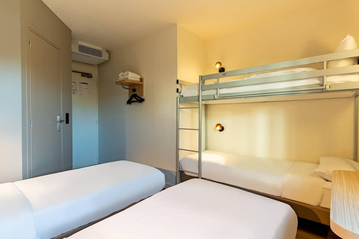 Una camera d'albergo con due letti singoli, un letto a castello, un armadio, un condizionatore e un tavolino con lampada.