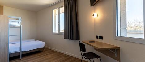Una habitación pequeña y bien iluminada con literas, un escritorio con silla, un televisor montado en la pared y una ventana con cortinas.