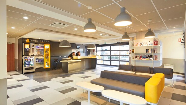 A modern lobby with a reception desk, vending machines, seating area, and a bookshelf. - Kyriad ECO - Marne-la-Vallée Bussy-Saint-Georges (Bussy-Saint-Georges)