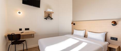 A hotel room with a bed, a desk with a chair, a TV mounted on the wall, and a wall-mounted light fixture.