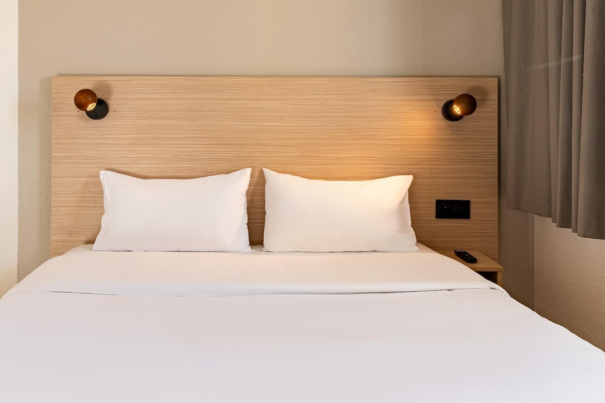 Un letto rifatto con due cuscini, una testiera in legno con due luci a parete e un comodino con un telecomando.