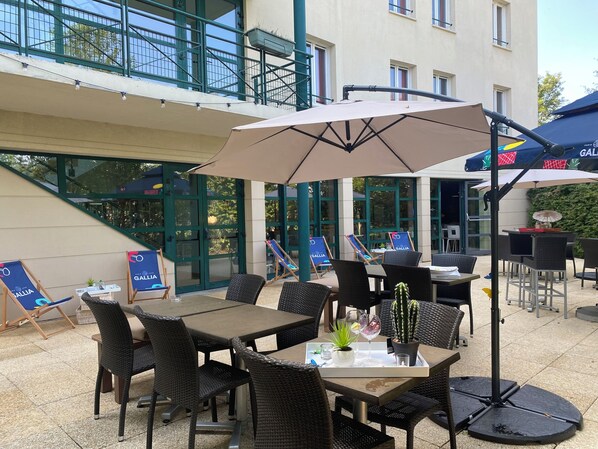 Un espace extérieur aménagé en terrasse, avec des meubles en osier, des tables et des chaises, dont certaines sont dotées de parasols.