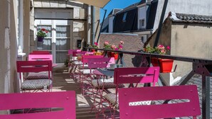 Una terraza exterior estrecha con sillas y mesas rosadas, plantas en macetas y vistas de edificios cercanos.