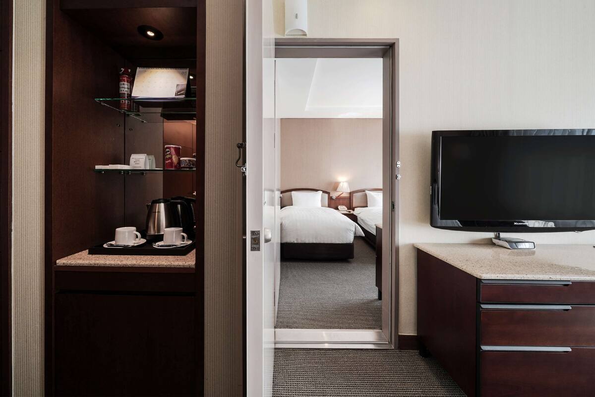Hotelkamer met een bed, nachtkastje, lamp, televisie en een kitchenette met een koffiezetapparaat en kopjes.