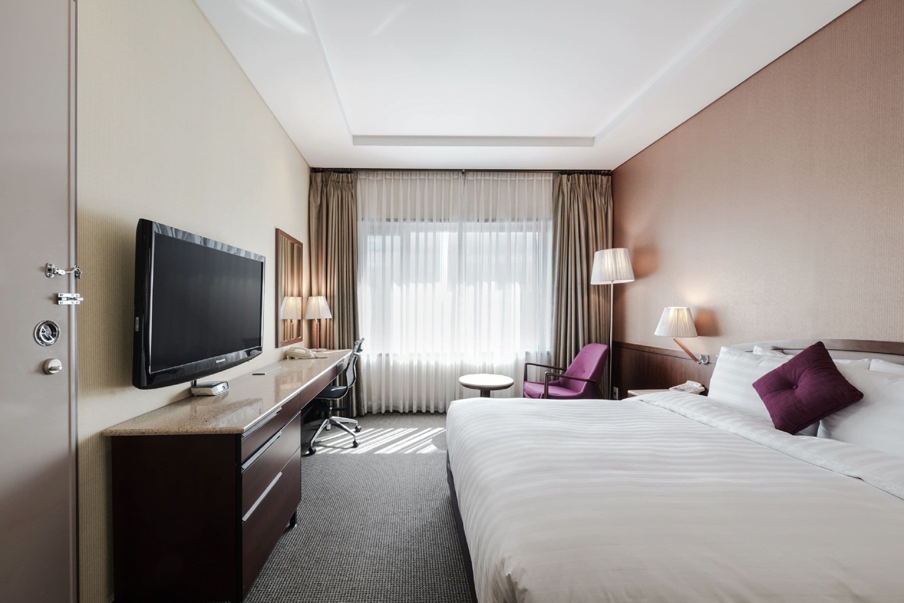A hotel room with a bed, a desk with a flat-screen TV, a chair, a small table, and a window with curtains.