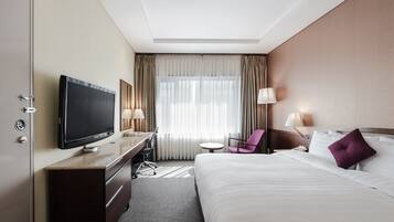 A hotel room with a bed, a desk with a flat-screen TV, a chair, a small table, and a window with curtains.